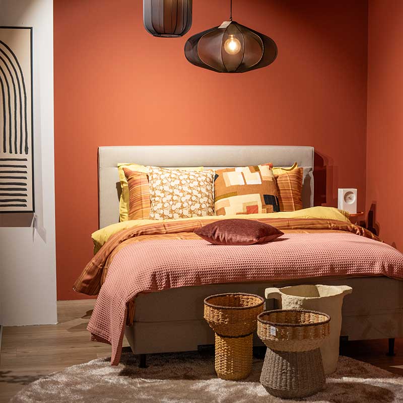 Slaapkamer met warme terracottamuren, kleurrijke kussens, designlamp en gevlochten manden voor een gezellige sfeer, geïnspireerd door de Blush ad Bold woontrend 2025.